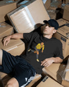 Man sitting among stacked cardboard boxes wearing a black hoodie with a graphic design.